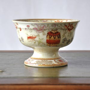 Könnte beinhalten: Eine weiße Keramik-Fußschale mit einem floralen Design in Rosa-, Rot- und Blautönen. Die Schale hat einen gewellten Rand und einen Fuß.