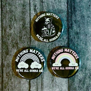 May include: Three black button badges with white text that reads "Nothing Matters We're All Gonna Die". The first badge features a black cat holding a skull. The second and third badges feature a rainbow with two white clouds with faces.