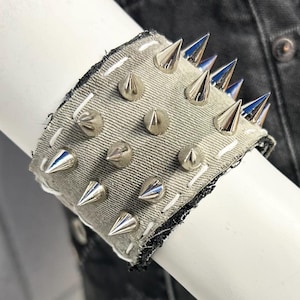 May include: A gray denim wrist cuff with silver-colored spikes. The cuff is trimmed with black fabric and white stitching. The cuff is on a mannequin arm, with black denim shorts in the background.
