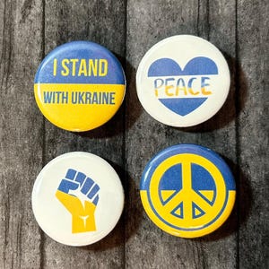 Puede incluir: Cuatro botones redondos amarillos y azules con diferentes mensajes y símbolos. Los botones dicen "Estoy con Ucrania", "Paz", y tienen un signo de paz y un puño en alto.