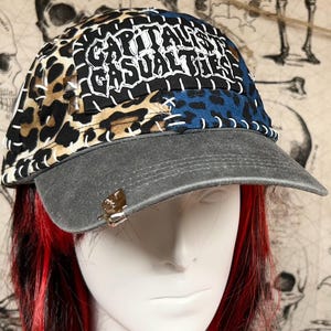 May include: A baseball cap with a gray brim and a patchwork design. The cap features leopard print, blue leopard print, and black fabric with the words "CAPITALIST CASUALTIES" in white. A metal accent is on the brim.