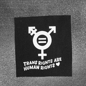 May include: Black fabric patch with a white transgender symbol and the text "Trans Rights Are Human Rights" with a white heart.