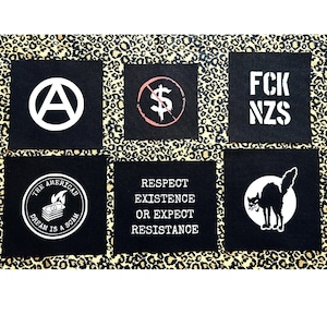 May include: Six black fabric patches with white and pink text and graphics. The patches include an anarchy symbol, a crossed-out dollar sign, the text "FCK NZS", a graphic of a burning stack of money with the text "The American Dream is a Scam", the text "Respect Existence or Expect Resistance", and a graphic of a black cat.
