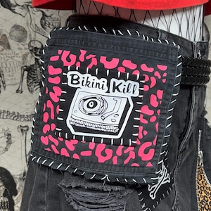 Puede incluir: Falda vaquera negra con detalles desgastados, con un parche con el texto "Bikini Kill" y un gráfico de tocadiscos. La falda tiene un acento de estampado de leopardo y está adornada con estrellas rosas y plateadas.