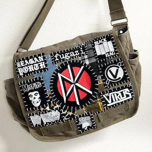 May include: An olive green canvas messenger bag featuring a collection of stitched fabric patches. The patches display various band names and logos including Reagan Youth, Fugazi, Black Flag, Misfits, The Virus, Bad Brains, and The Exploited.