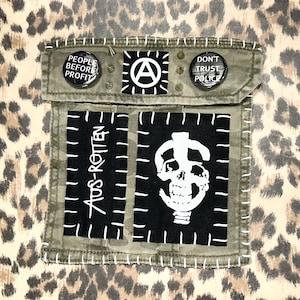 May include: A camouflage fabric pocket featuring two black rectangular patches with white screen-printed designs, one reading "AUS-ROTTEN" and the other a skull. Two circular buttons read "PEOPLE BEFORE PROFIT" and "DON'T TRUST POLICE" with an anarchy symbol.