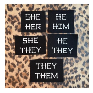 May include: Five black fabric patches with white text. The patches read "She Her", "He Him", "She They", "He They", and "They Them".