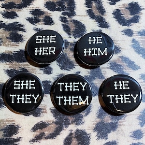 May include: Five black button badges with white text. The badges read "She Her", "He Him", "She They", "They Them", and "He They".
