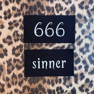 May include: Two black fabric patches with white text. The top patch has the number 666. The bottom patch says "sinner".