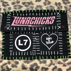 May include: A black fabric patch featuring the pink text LUNACHICKS at the top. Below, a white circle contains the text L7, and a white heart outline contains the text BABES IN TOYLAND. The patch has white stitched borders and neon green edge stitching.