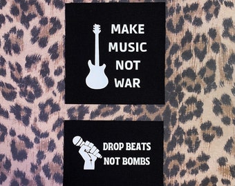 May include: Two black fabric patches with white text and graphics. The top patch features a white electric guitar and the text "MAKE MUSIC NOT WAR". The bottom patch features a white microphone and fist and the text "DROP BEATS NOT BOMBS".