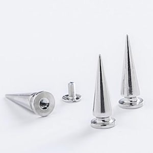 Può includere: Una collezione di borchie e elementi di fissaggio a forma di cono in metallo color argento. Le borchie hanno una punta e una base circolare. Una borchia è cilindrica con un'estremità piatta. Gli oggetti sono disposti su una superficie bianca.