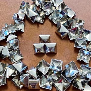 May include: A collection of shiny, silver-colored metal pyramid studs arranged on a brown surface. The studs are square-based pyramids with a reflective surface, creating a geometric pattern. The arrangement suggests a craft or DIY project.