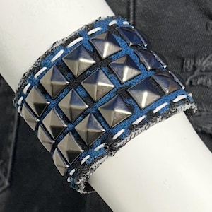 Puede incluir: Una pulsera de puño de mezclilla azul con filas de tachuelas piramidales plateadas. La pulsera tiene costuras blancas en los bordes y se usa en el brazo de un maniquí. Un accesorio atrevido.