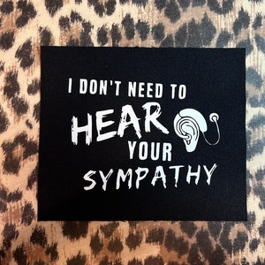 Puede incluir: Parche de tela negro con texto blanco que dice "No necesito oír tu SIMPATÍA" con un dibujo blanco de una oreja y un audífono.