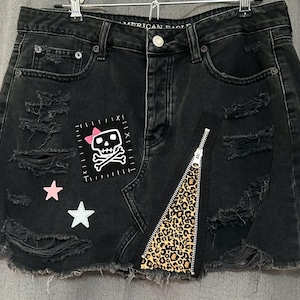 Può includere: Gonna di jeans nera con dettagli consumati, con una toppa a forma di teschio e ossa incrociate, un fiocco rosa, un pannello di tessuto con stampa leopardata e un dettaglio di cerniera.