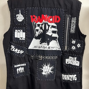 May include: A black sleeveless denim vest featuring numerous white and red band patches hand-stitched onto the back. Patches include Rancid, Black Flag, The Clash, Ramones, Bikini Kill, Sex Pistols, and Danzig, along with a central belt and zipper.