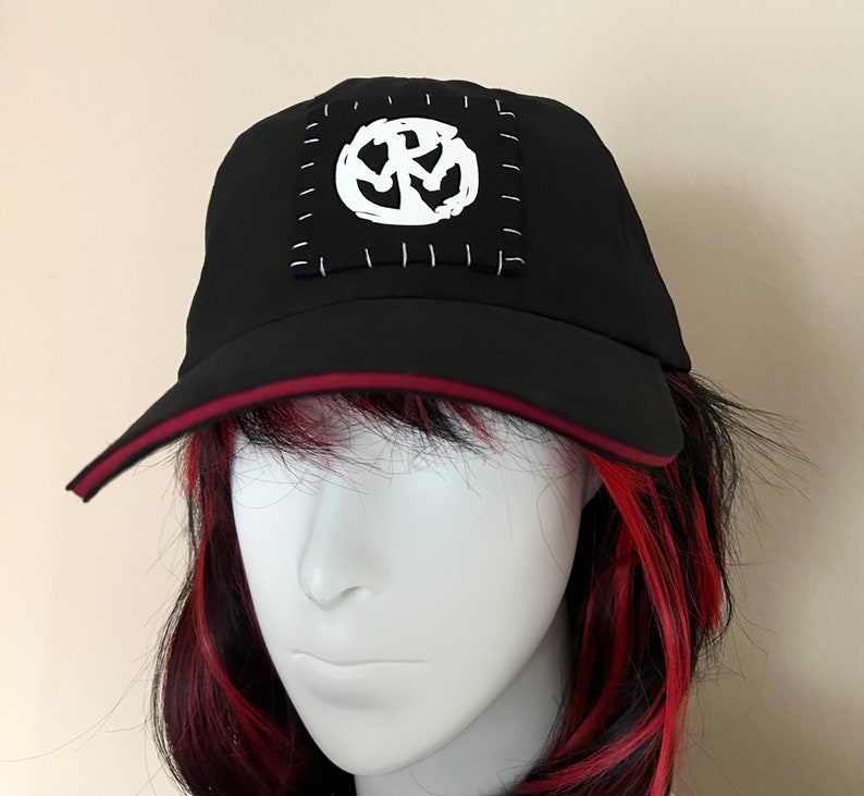 May include: Black baseball cap with a white graphic patch. The cap features a red trim along the brim and a white stitched patch with a unique design. The hat is displayed on a mannequin head.