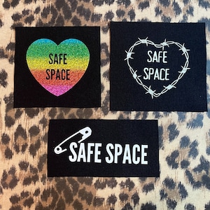 Könnte beinhalten: Drei schwarze Stoff-Patches mit weißem Text und Designs. Der erste Patch hat ein Regenbogen-Glitzerherz mit dem Text "SAFE SPACE". Der zweite Patch hat ein weißes Stacheldraht-Herz mit dem Text "SAFE SPACE". Der dritte Patch hat eine weiße Sicherheitsnadel und den Text "SAFE SPACE".
