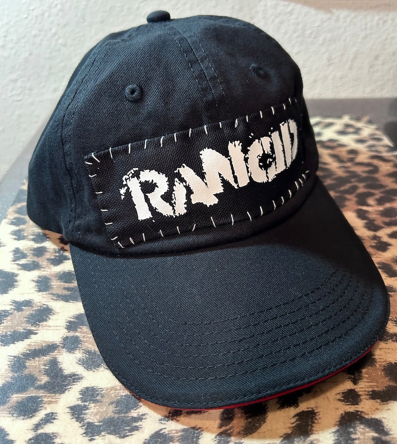 May include: Black baseball cap with a curved brim and a patch featuring the word "RANCID" in white, stitched onto the front. The cap has a red trim along the brim's edge.