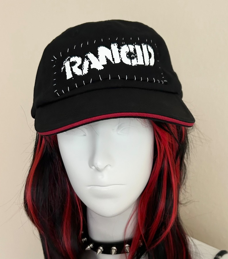 May include: Black baseball cap with a red brim and the word "RANCID" in white block letters on a patch. The patch is stitched onto the cap with white thread. The cap is displayed on a mannequin head.