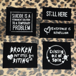 May include: Five black patches with white text. The patches read: "SUICIDE IS A PERMANENT SOLUTION TO A TEMPORARY PROBLEM", "STILL HERE", "EXISTENCE IS REBELLION", "BROKEN BUT STILL BITING", and "Existing out of Spite".