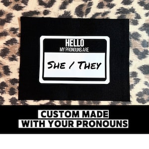 May include: Black fabric patch with a white name tag that says "Hello, my pronouns are she/they". The text "Custom made with your pronouns" is printed below the name tag.