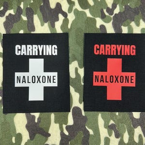 Op de afbeelding: Twee zwarte vierkante patches met witte tekst en een wit kruis, en een rood kruis. De tekst op de patches luidt "CARRYING NALOXONE". De achtergrond is een camouflagepatroon in tinten groen, bruin en beige.