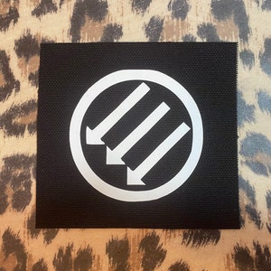 May include: A square, black patch featuring a white circle with three downward-pointing arrows inside. The patch is set against a leopard print background.