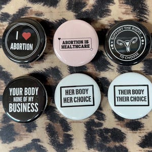 May include: Six black and white button badges with pro-choice slogans. The slogans are "I love abortion", "Abortion is healthcare", "Support the right to choose", "Your body none of my business", "Her body her choice", and "Their body their choice".