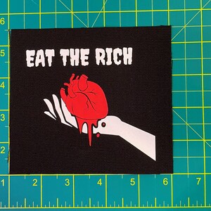 May include: Black square patch with the text "EAT THE RICH" in white block letters. A red anatomical heart with dripping blood is held by a white hand. The patch measures approximately 4 inches by 4 inches.