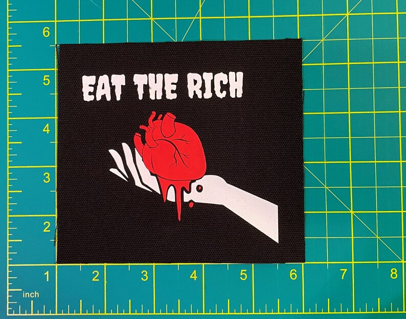 May include: Black square patch with the text "EAT THE RICH" in white block letters. A red anatomical heart with dripping blood is held by a white hand. The patch measures approximately 4 inches by 4 inches.