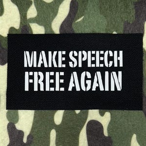 Puede incluir: Un parche rectangular negro con el texto blanco "MAKE SPEECH FREE AGAIN" sobre un fondo de camuflaje. El diseño de camuflaje presenta tonos de verde, marrón y beige.