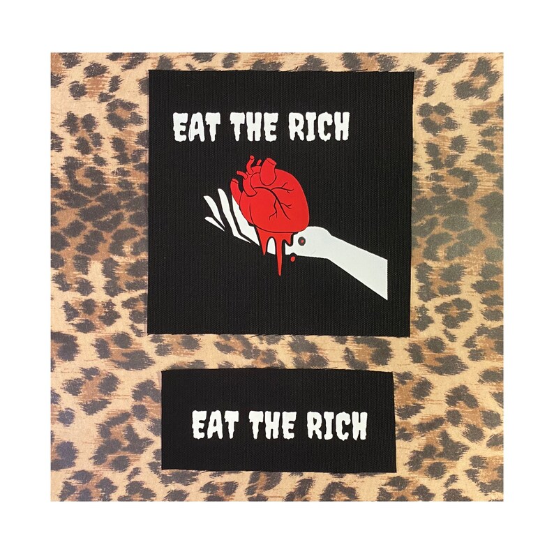 May include: Two black patches with white text and a graphic design. The larger patch features the text "EAT THE RICH" above a red heart dripping blood, held by a white hand. The smaller patch below repeats the text "EAT THE RICH".