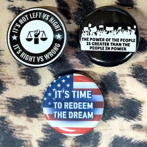 May include: Three round button pins with political messages. The first pin is black and white with the text "It's not left vs right, it's right vs wrong" and a scale of justice. The second pin is black with white text that reads "The power of the people is greater than the people in power". The third pin has an American flag design with the text "It's time to redeem the dream".