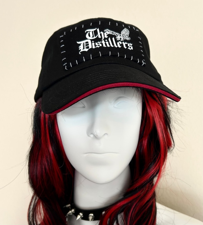May include: Black baseball cap with a red brim, featuring the text "The Distillers" in white gothic lettering. The patch is stitched onto the cap with white thread. A skull graphic is also present on the patch.