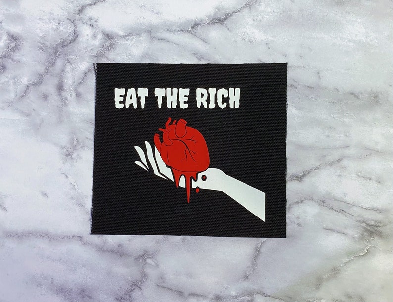 May include: Black square patch with the text "EAT THE RICH" in white. A red heart with dripping blood is held in a white hand. The patch is likely for clothing or accessories.