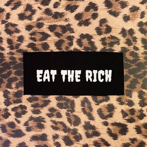 May include: A black rectangular patch with the white text "EAT THE RICH" is centered on a background with a brown and black leopard print pattern.