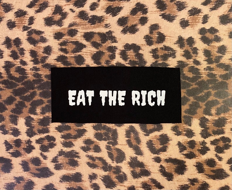 May include: A black rectangular patch with the white text "EAT THE RICH" is centered on a background with a brown and black leopard print pattern.