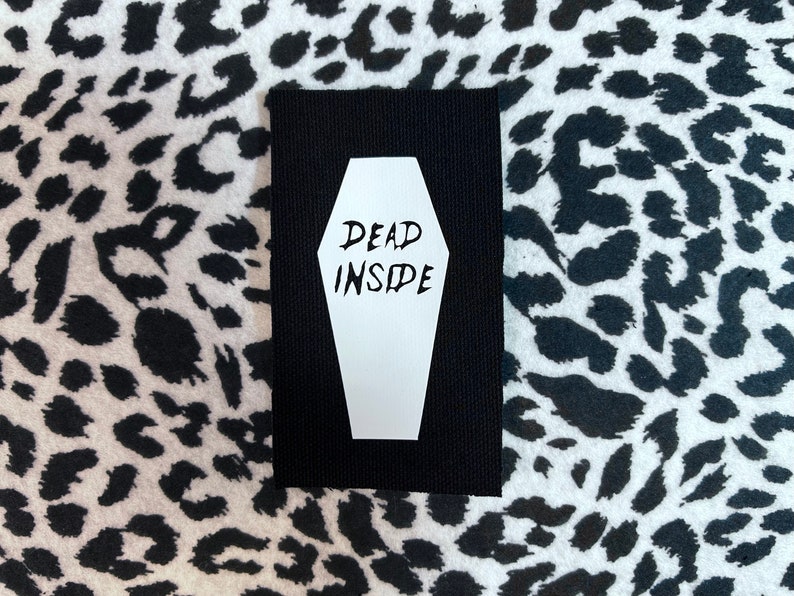May include: Black fabric patch with a white coffin shape and the text "DEAD INSIDE" inside the coffin.