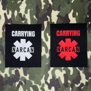 May include: Two black patches with white and red text and a star symbol. The text on both patches reads "CARRYING NARCAN".