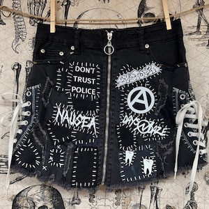 May include: Black denim skirt with a zipper closure and white stitched patches. The skirt features the phrases "DON'T TRUST POLICE" and "NAUSEA", along with other graphics and text. White laces are on the sides.