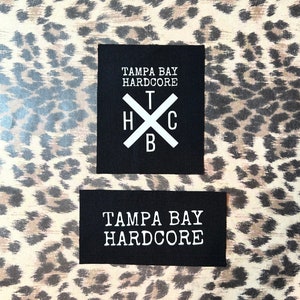 May include: Two black fabric patches with white text. The top patch has the text "Tampa Bay Hardcore" and a white X with the letters "T", "H", "C", and "B" inside. The bottom patch has the text "Tampa Bay Hardcore".