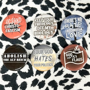 May include: A collection of six round buttons with various anti-fascist and anti-hate messages. The buttons feature bold text on colorful backgrounds, including phrases like "Smash Christo-Fascism" and "God Hates Flags."
