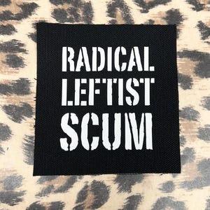 May include: Black square patch featuring the words "RADICAL. LEFTIST. SCUM" in white stencil lettering. The patch is displayed on a leopard print background.