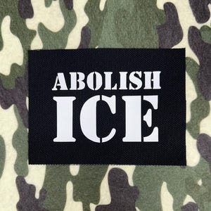May include: A black rectangular patch with the words "ABOLISH ICE" in bold white capital letters. The patch is set against a camouflage pattern background in shades of green, brown, and beige.