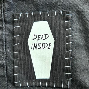 May include: Black denim jeans with a white patch sewn on. The patch is shaped like a coffin and has the words "DEAD INSIDE" written on it.