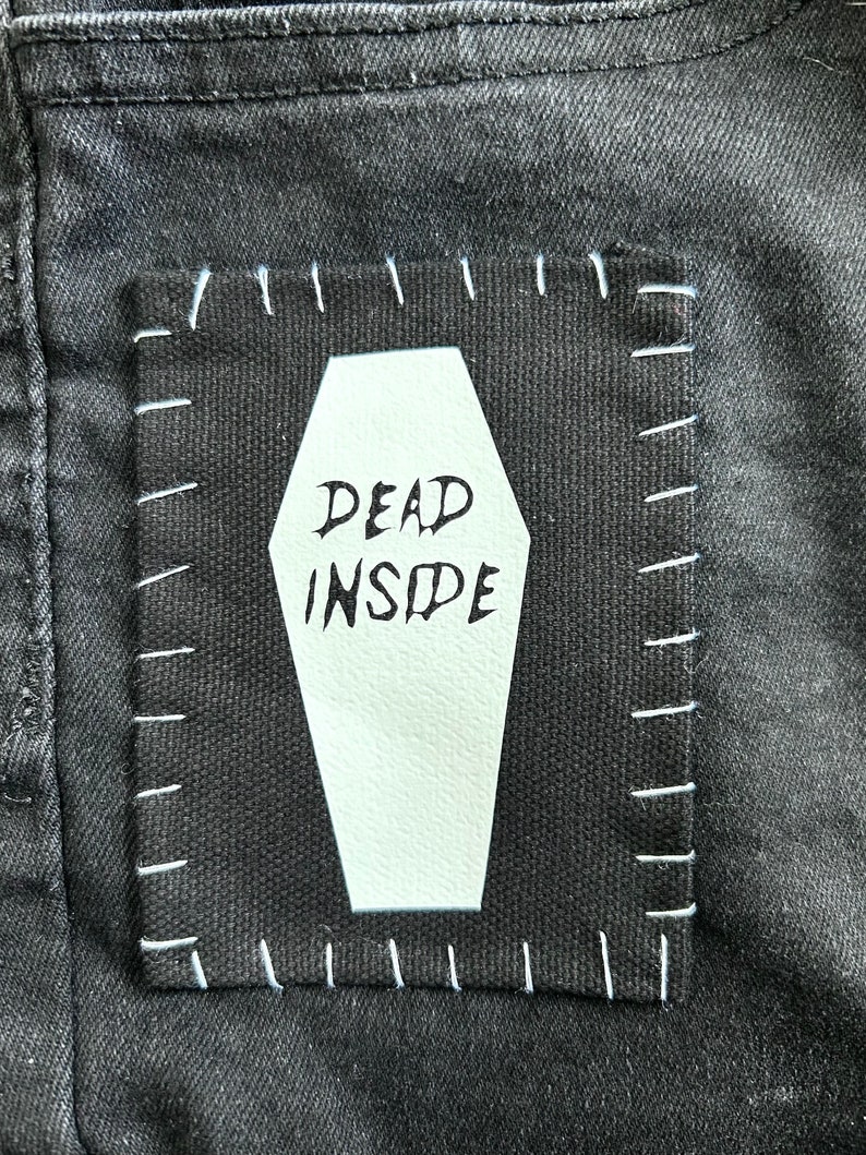 May include: Black denim jeans with a white patch sewn on. The patch is shaped like a coffin and has the words "DEAD INSIDE" written on it.