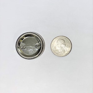 May include: The back of a silver button with a pin and a quarter for size comparison.
