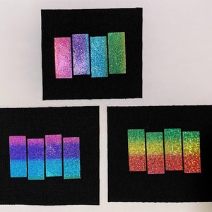 May include: Three black fabric patches with different colored iridescent stripes. The top patch has pink, purple, and blue stripes. The middle patch has pink, purple, and blue stripes. The bottom patch has green, yellow, and red stripes.